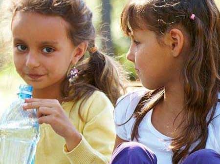 Two young girls sitting outdoors, one holding a plastic water bottle and looking at the other. - Olive Oil Times