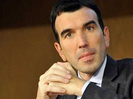 A man in a dark suit with short black hair, sitting with his hands clasped in front of him. - Olive Oil Times