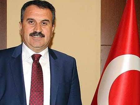 A man in a suit standing next to a Turkish flag, with a neutral expression and a mustache. - Olive Oil Times