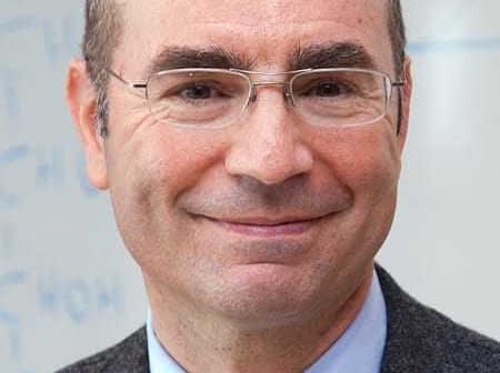 A man wearing glasses and a gray suit, smiling at the camera with a neutral background. - Olive Oil Times