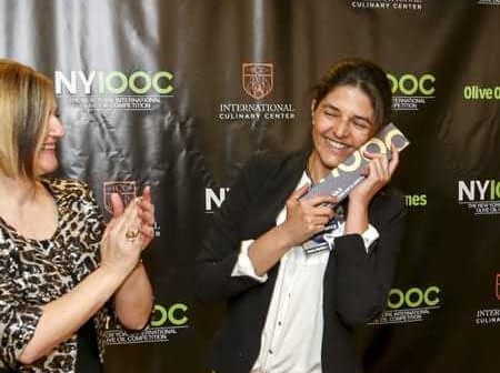 A woman smiling while holding an award at an event with a backdrop featuring Olive Oil Times and NYIOOC logos. - Olive Oil Times