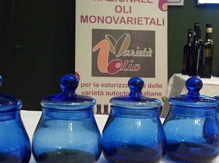 Four blue glass jars arranged in a row at an olive oil event with a promotional banner in the background. - Olive Oil Times