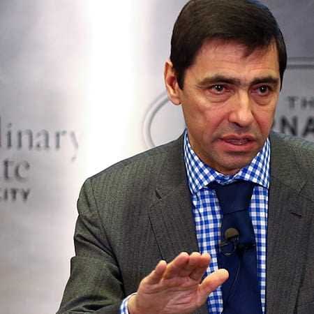 Man in a gray suit with a blue checkered shirt speaking during a presentation. - Olive Oil Times