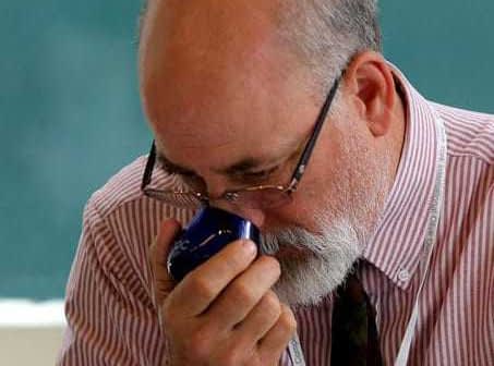 A man with glasses and a beard smelling a blue container in a classroom setting. - Olive Oil Times