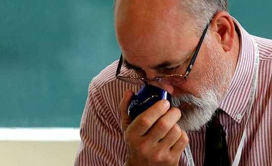 A man with glasses and a beard holding a blue object close to his face in a classroom setting. - Olive Oil Times