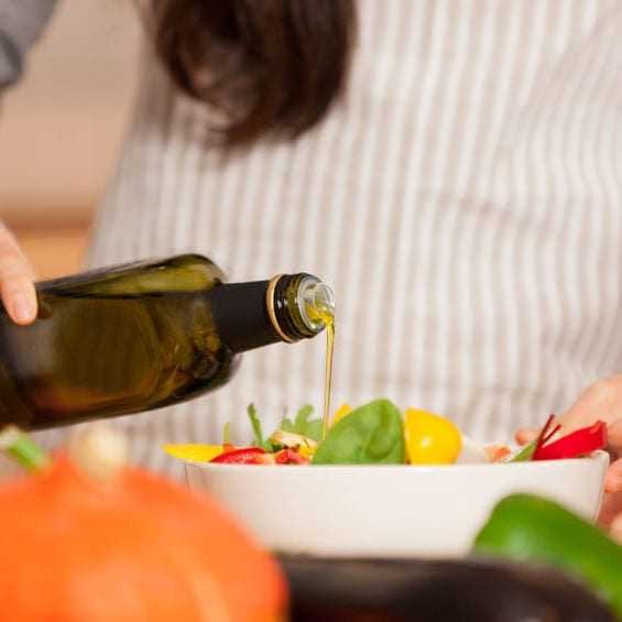 Una persona vierte aceite de oliva de una botella en un recipiente con ingredientes frescos para ensalada. Olive Oil Times