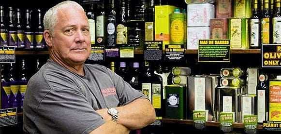 Man with gray hair standing with arms crossed in front of olive oil bottles on shelves. - Olive Oil Times