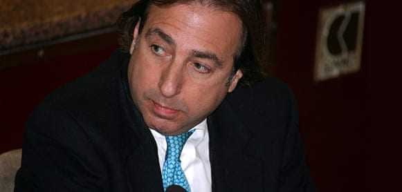 Man wearing a black suit and a blue tie, looking to the side during a discussion. - Olive Oil Times