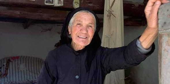 Elderly woman wearing a black dress and headscarf, smiling while leaning against a wall. - Olive Oil Times