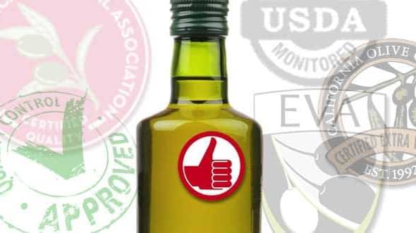 Green glass bottle of olive oil featuring a thumbs-up symbol and certification seals in the background. - Olive Oil Times