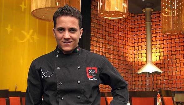 Chef wearing a black culinary uniform with a logo, standing in a restaurant setting. - Olive Oil Times