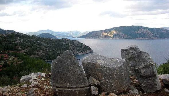 Three large stone fragments positioned near the coastline with a view of the sea and mountains in the background. - Olive Oil Times