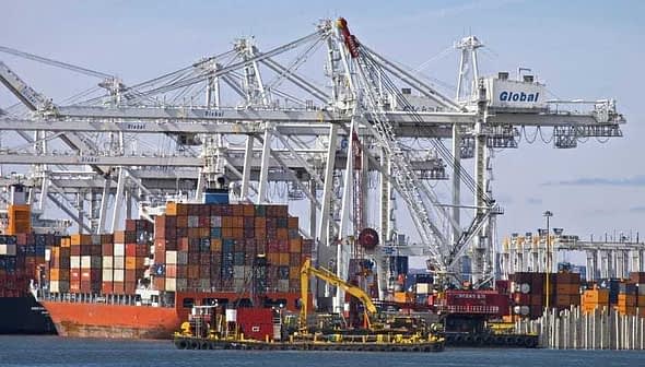 Cargo ship docked at a port with stacked shipping containers and cranes in the background. - Olive Oil Times