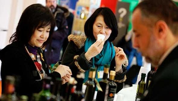 Two women sampling olive oil at a trade show, one holding a tasting cup and the other observing. - Olive Oil Times