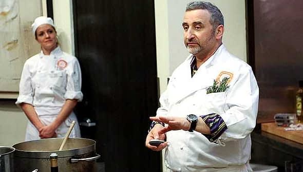 Chef in a white coat demonstrating cooking techniques with a pot in front of him and a female chef in the background. - Olive Oil Times