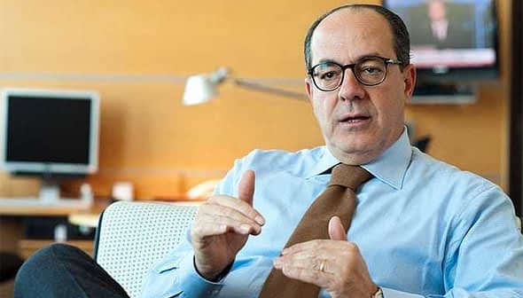 Businessman wearing a light blue shirt and glasses, gesturing while seated in an office environment. - Olive Oil Times