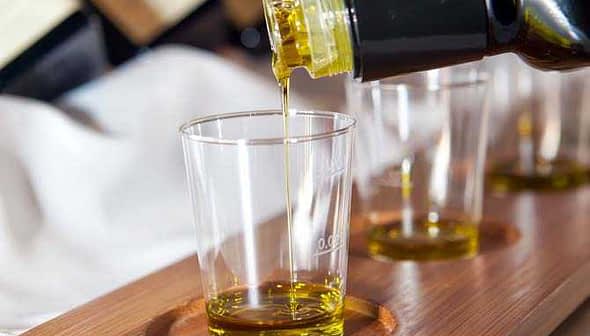 Olive oil being poured from a bottle into a clear glass sample cup on a wooden tray. - Olive Oil Times
