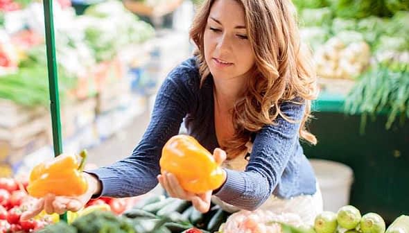 Woman holding two yellow bell peppers while shopping at a market filled with various vegetables. - Olive Oil Times
