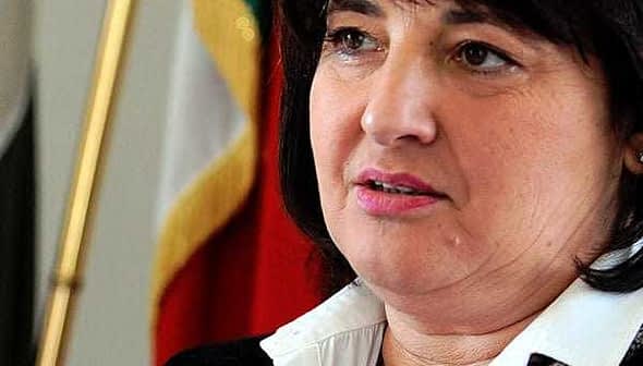 Woman with dark hair and a white shirt speaking in front of flags. - Olive Oil Times