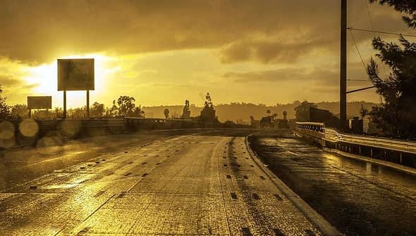 A wet road with reflective surfaces during sunset, featuring distant trees and signage. - Olive Oil Times