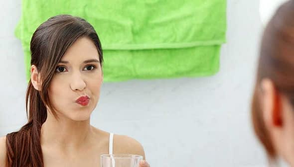 A woman rinsing her mouth with water while looking in a mirror in a bathroom. - Olive Oil Times