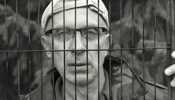 A man wearing glasses and a cap, looking through metal bars with a serious expression. - Olive Oil Times