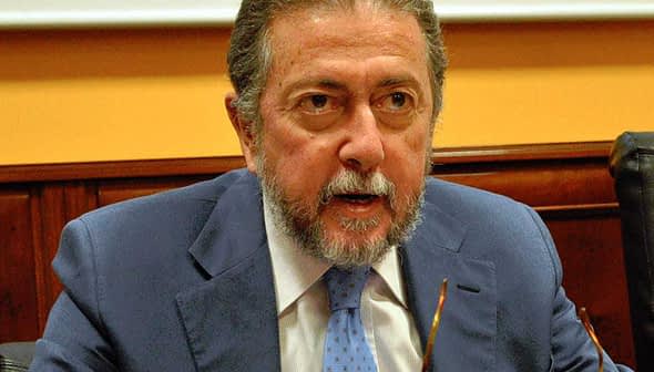 Man with gray hair and beard wearing a blue suit speaking at a conference table. - Olive Oil Times