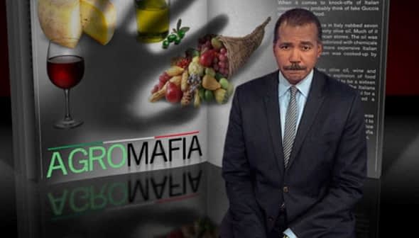 A man in a suit standing next to a graphic titled 'Agro Mafia' with food items and a glass of wine. - Olive Oil Times