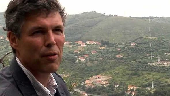A man in a suit standing in front of an olive grove on a hillside. - Olive Oil Times