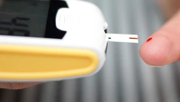 Person using a blood glucose meter with a test strip inserted and a drop of blood on their fingertip. - Olive Oil Times