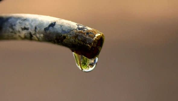 Close-up of a water droplet hanging from the tip of a faucet spout. - Olive Oil Times