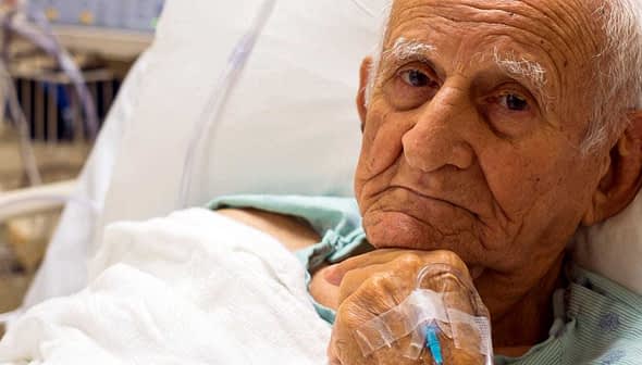 Elderly man lying in a hospital bed with an intravenous line in his hand. - Olive Oil Times