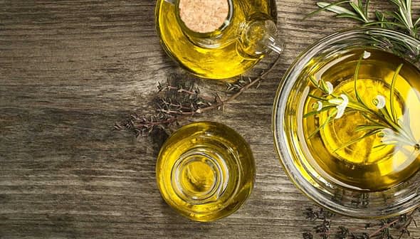 Glass bowl and bottle containing olive oil with herbs on a wooden surface. - Olive Oil Times