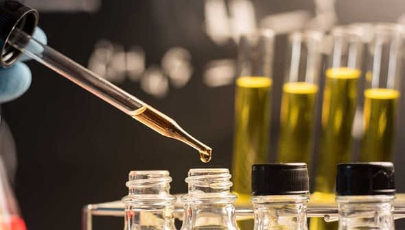 A hand with a blue glove holding a pipette above several test tubes containing yellow liquid. - Olive Oil Times