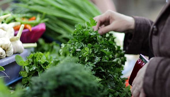 Person selecting fresh parsley from a display of herbs and vegetables at a market. - Olive Oil Times