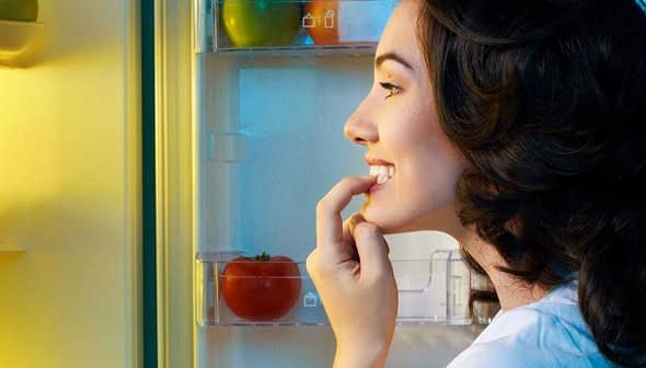 A woman with dark hair smiling while looking into an open refrigerator filled with food items. - Olive Oil Times
