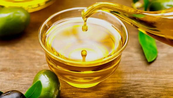 A glass bowl filled with olive oil with a stream pouring from a vessel and olives nearby. - Olive Oil Times
