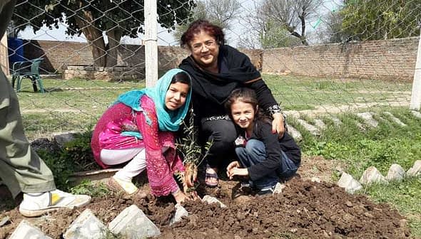 A woman and two children planting a small tree in a garden area with a fence in the background. - Olive Oil Times