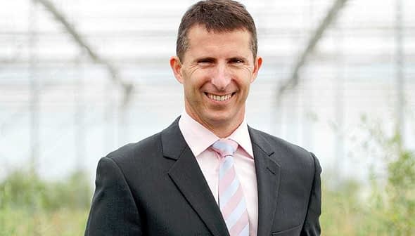 Man wearing a dark suit and light blue striped tie, smiling in a greenhouse setting. - Olive Oil Times