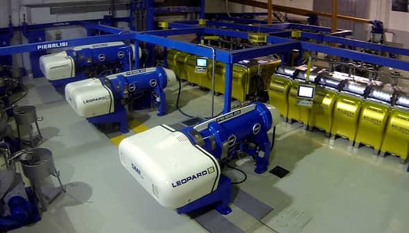 Industrial olive oil production equipment with blue and yellow machines in a processing facility. - Olive Oil Times