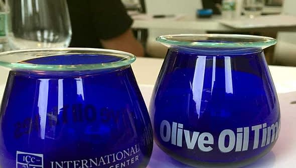 Two blue glass cups labeled 'International Culinary Center' and 'Olive Oil Times' on a table. - Olive Oil Times