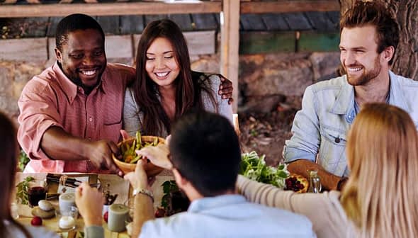 A diverse group of people sitting around a table sharing food in an outdoor setting. - Olive Oil Times
