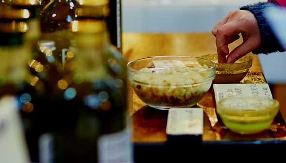 A person dipping bread into a bowl of olive oil for tasting at a display. - Olive Oil Times