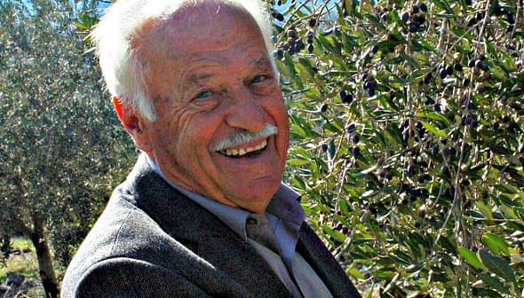 Elderly man smiling while standing next to olive trees with ripe olives. - Olive Oil Times