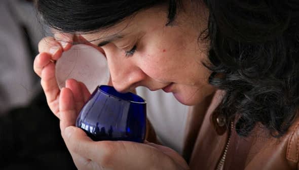 Woman with dark hair holding a blue glass container close to her face, smelling the contents. - Olive Oil Times