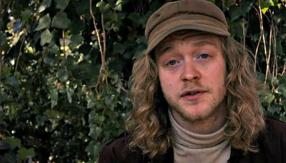 A man with long, curly hair wearing a beige cap and a turtleneck sweater, sitting outdoors. - Olive Oil Times