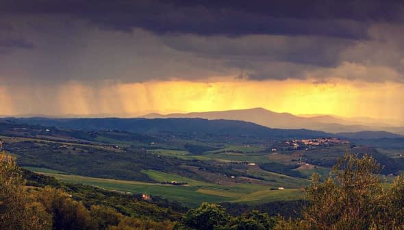Panoramic view of rolling hills with dark clouds and sunlight breaking through in the distance. - Olive Oil Times