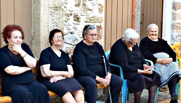 Six elderly women dressed in black and one in pink sitting on a bench outside a building. - Olive Oil Times