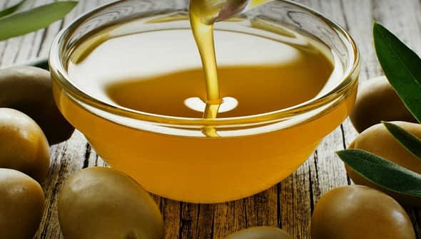 A bowl of olive oil being poured, surrounded by fresh green olives on a wooden surface. - Olive Oil Times