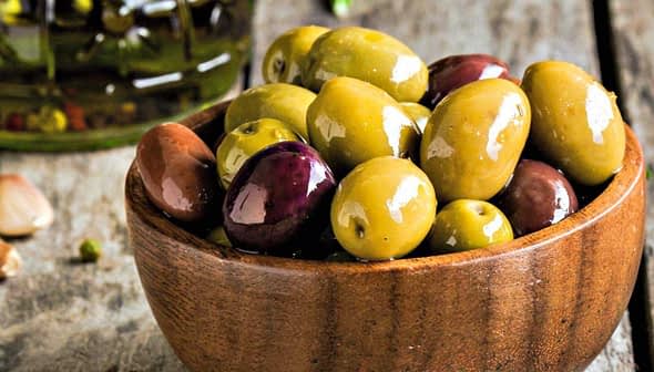 A wooden bowl filled with various types of olives including green and black varieties. - Olive Oil Times